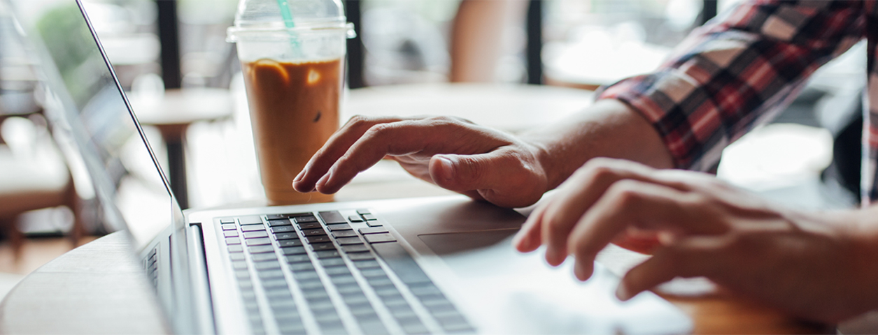 An online student's hands are hovering over a laptop. An iced coffee also sits on the table.
